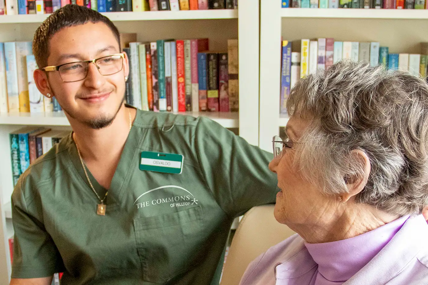 A staff member at The Commons of Hilltop smiles while sitting beside an older woman in a library.